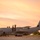A U.S. military C-130 Hercules turboprop transport aircraft, marked with tail code 'QD' and serial number '9532', is parked on a tarmac at what appears to be a tropical airfield during sunset or sunrise. Several personnel in flight suits or military uniforms are visible near the aircraft's forward entry door, likely conducting pre-flight or post-flight activities. Palm trees are visible in the background, suggesting a Pacific or tropical island location.