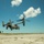 A military tiltrotor aircraft, identifiable as a Bell Boeing V-22 Osprey, is captured in low-altitude flight just above a runway or airstrip in what appears to be a tropical coastal location. The rotors are in helicopter mode (tilted upward), suggesting the aircraft is in a hovering or slow-speed flight phase, likely landing or taking off. At least two additional Osprey aircraft are visible in the background, indicating a multi-aircraft operation.