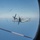 The image is taken through a porthole/oval window of one aircraft looking out at a V-22 Osprey tiltrotor aircraft flying in formation nearby. The Osprey, marked with the number '05' on its fuselage, appears to be a U.S. Marine Corps MV-22 Osprey with rotors in airplane mode. A refueling drogue line with orange-and-white banding is visible in the foreground, suggesting an aerial refueling operation may be in progress.