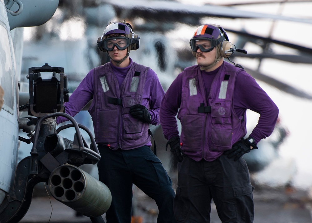 Two military personnel wearing purple jerseys and safety gear stand on what appears to be an aircraft carrier flight deck. They are positioned next to a military helicopter equipped with a rocket pod launcher. The purple jerseys identify them as aviation ordnancemen or fuel handlers in U.S. Navy flight deck operations.