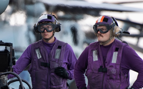 Two military personnel wearing purple jerseys and safety gear stand on what appears to be an aircraft carrier flight deck. They are positioned next to a military helicopter equipped with a rocket pod launcher. The purple jerseys identify them as aviation ordnancemen or fuel handlers in U.S. Navy flight deck operations.