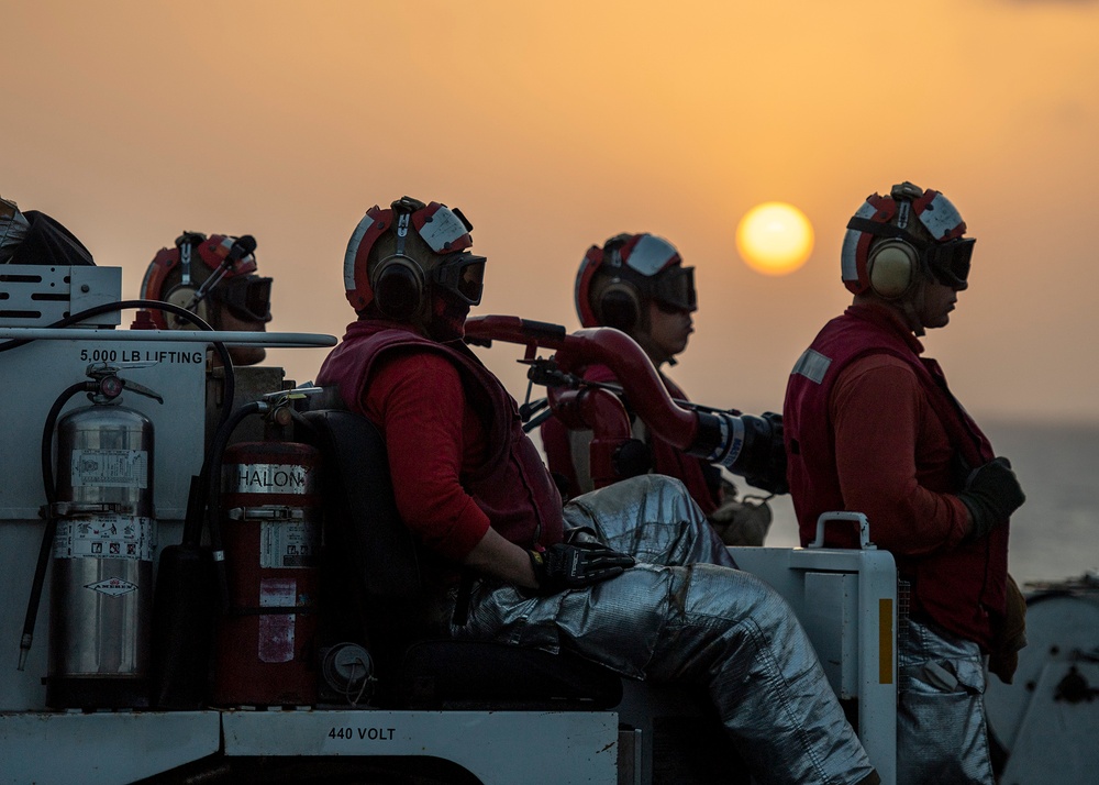 A group of naval firefighting crew members dressed in red jerseys, protective helmets with ear protection, and silver fire-resistant trousers are positioned on what appears to be a flight deck vehicle at sunset. The personnel are equipped with fire suppression equipment including Halon fire extinguishers and appear to be on standby duty. The dramatic orange sunset over open water provides a striking backdrop to this military operational scene.