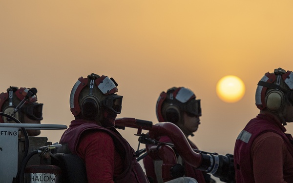 A group of naval firefighting crew members dressed in red jerseys, protective helmets with ear protection, and silver fire-resistant trousers are positioned on what appears to be a flight deck vehicle at sunset. The personnel are equipped with fire suppression equipment including Halon fire extinguishers and appear to be on standby duty. The dramatic orange sunset over open water provides a striking backdrop to this military operational scene.