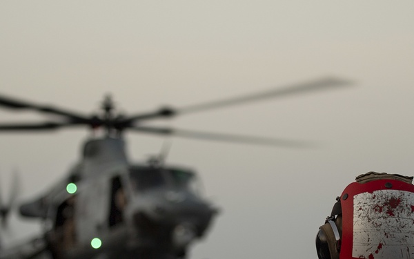 A military helicopter, appearing to be a UH-1Y Venom, is hovering or landing on a naval vessel's flight deck at dusk or dawn. Deck crew members in colored safety vests and helmets are directing the aircraft using hand signals. The foreground shows a crew member in a red vest and helmet observing the operation.