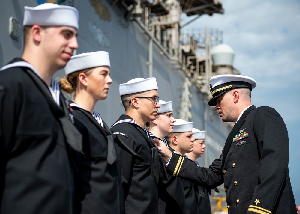 A U.S. Navy officer in dress blues with gold insignia and ribbons is inspecting or presenting awards to a line of enlisted sailors in service dress blue uniforms with white Dixie cup covers. The ceremony appears to be taking place on or near a naval vessel, visible in the background. The officer is closely examining or pinning something on one of the sailors standing at attention.