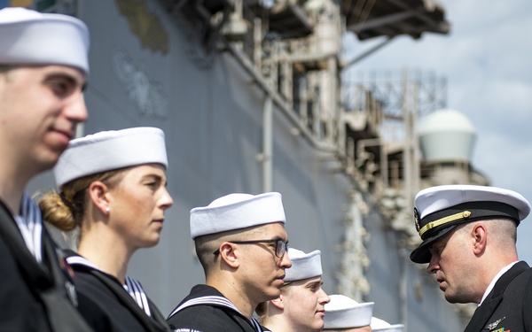A U.S. Navy officer in dress blues with gold insignia and ribbons is inspecting or presenting awards to a line of enlisted sailors in service dress blue uniforms with white Dixie cup covers. The ceremony appears to be taking place on or near a naval vessel, visible in the background. The officer is closely examining or pinning something on one of the sailors standing at attention.