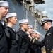 A U.S. Navy officer in dress blues with gold insignia and ribbons is inspecting or presenting awards to a line of enlisted sailors in service dress blue uniforms with white Dixie cup covers. The ceremony appears to be taking place on or near a naval vessel, visible in the background. The officer is closely examining or pinning something on one of the sailors standing at attention.