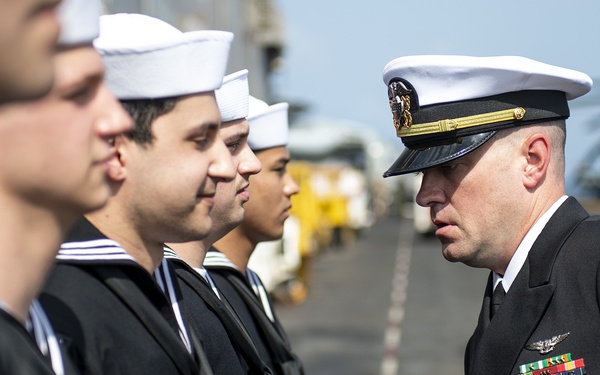 A senior U.S. Navy officer in dress blues with a combination cover and multiple service ribbons is conducting an inspection of enlisted sailors standing in formation. The sailors are wearing standard Navy dress blue uniforms with white Dixie cup covers. The setting appears to be the flight deck of a naval vessel, with aircraft and deck equipment visible in the background.