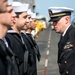 A senior U.S. Navy officer in dress blues with a combination cover and multiple service ribbons is conducting an inspection of enlisted sailors standing in formation. The sailors are wearing standard Navy dress blue uniforms with white Dixie cup covers. The setting appears to be the flight deck of a naval vessel, with aircraft and deck equipment visible in the background.
