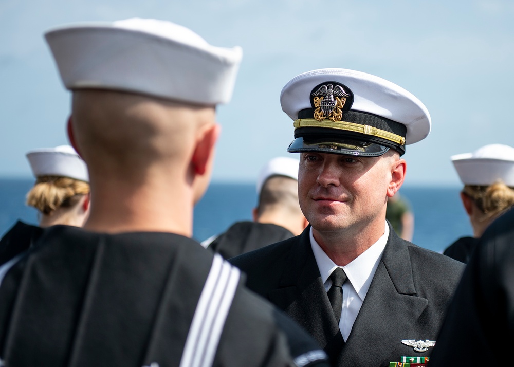 A U.S. Navy officer in Service Dress Blue uniform with a white combination cover is facing enlisted sailors also in Service Dress Blue uniforms. The officer appears to be conducting an inspection or formal review, standing face-to-face with a sailor whose back is to the camera. The setting appears to be outdoors near a body of water, suggesting a shipboard or pier-side ceremony.