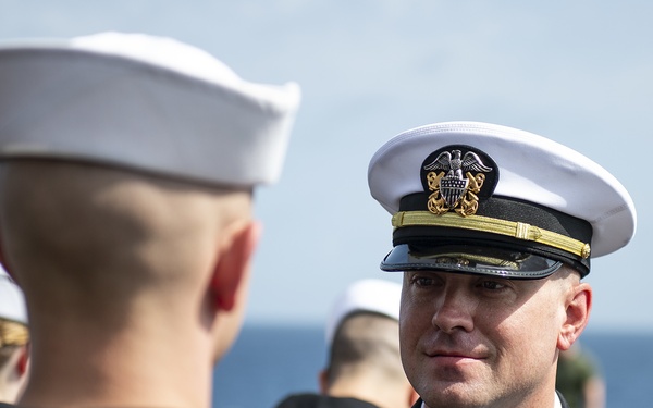 A U.S. Navy officer in Service Dress Blue uniform with a white combination cover is facing enlisted sailors also in Service Dress Blue uniforms. The officer appears to be conducting an inspection or formal review, standing face-to-face with a sailor whose back is to the camera. The setting appears to be outdoors near a body of water, suggesting a shipboard or pier-side ceremony.