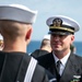 A U.S. Navy officer in Service Dress Blue uniform with a white combination cover is facing enlisted sailors also in Service Dress Blue uniforms. The officer appears to be conducting an inspection or formal review, standing face-to-face with a sailor whose back is to the camera. The setting appears to be outdoors near a body of water, suggesting a shipboard or pier-side ceremony.