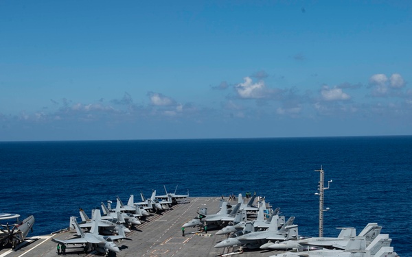 An aerial view of a U.S. Navy aircraft carrier flight deck at sea, densely packed with multiple fixed-wing aircraft including F/A-18 Super Hornets and at least one E-2 Hawkeye airborne early warning aircraft. The carrier is underway in open blue ocean under clear skies. Deck crew personnel are visible at various positions on the flight deck.