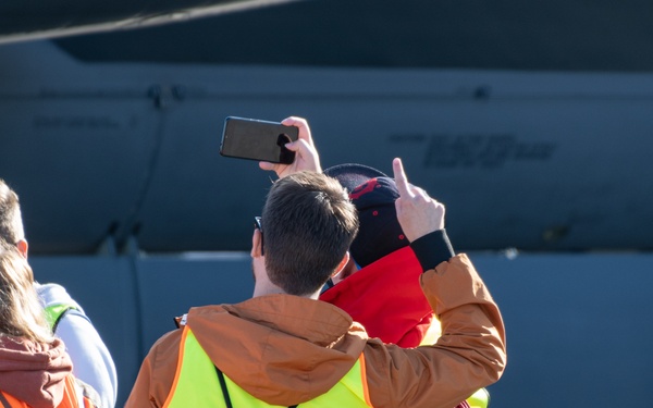 A group of people wearing high-visibility safety vests are standing on what appears to be an airfield or tarmac, looking at a large aircraft. One person in the center is holding up a smartphone to take a photo of the aircraft. The group appears to be on an organized tour or visit, as indicated by their matching safety vests.