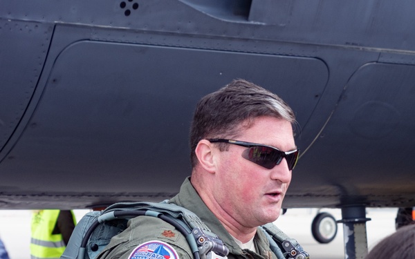 A military pilot in an olive green flight suit is speaking to a small group of people, likely at an airshow or public aviation event. He is standing beneath the fuselage of a dark-colored military aircraft. He wears sunglasses, a flight harness, and several patches on his uniform.