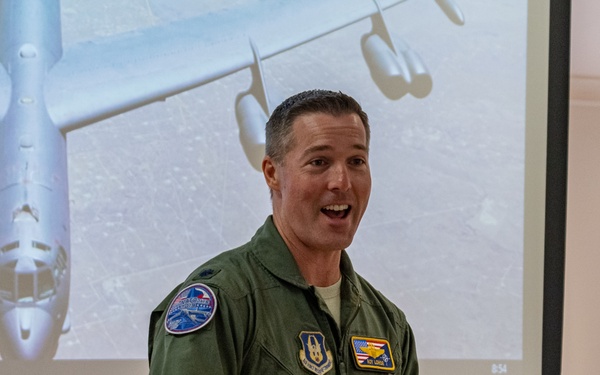A military aviator in a green flight suit is giving a presentation or briefing in what appears to be a classroom or conference room. Behind him, a projected image shows an aerial photograph of a large military aircraft, likely a bomber or tanker, taken from above. The individual appears engaged and animated while speaking.