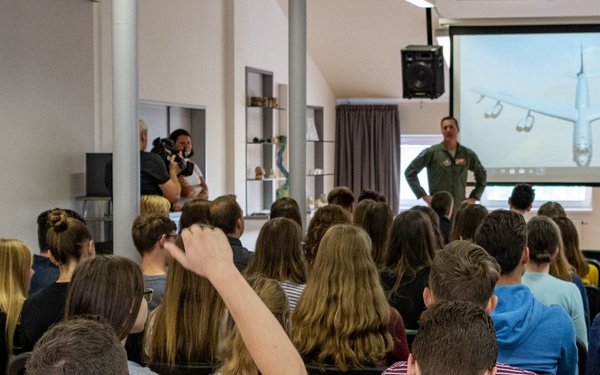 Two military personnel in flight suits are presenting to a large group of students or young adults in a classroom or lecture hall setting. A projection screen behind them displays what appears to be a military aircraft viewed from below. One audience member has their hand raised, suggesting an interactive Q&amp;A session.