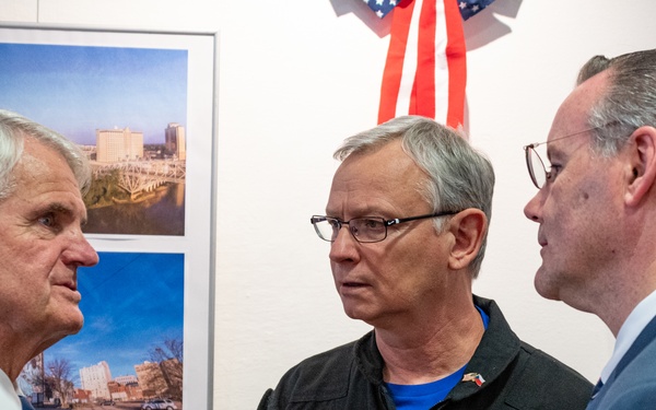 Three men are engaged in conversation at what appears to be a formal or semi-formal indoor event. The central figure is wearing a flight jacket with patches identifying him as 'Joe Jones' from the 'Trojan Phlyers T-28 Flight Demonstration Team.' An American flag is visible in the background along with framed photographs on the wall.