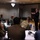 Taking the Lead to Connect A civilian presenter in a dark suit and red tie is speaking to a room of military personnel wearing camouflage uniforms. The setting appears to be a conference or workshop held in a banquet-style room, with attendees seated at round tables. A whiteboard or easel and a sign on one of the tables suggest a structured training or briefing event.