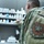 DHA to provide integrated health and readiness A U.S. Air Force Space Command service member is shown from behind, examining medication bottles on pharmacy shelving. The individual is wearing an Operational Camouflage Pattern (OCP) uniform with a clearly visible 'Air Force Space Command' shoulder patch. The setting appears to be a military pharmacy or medical supply room.