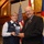 Never Forget, Never Lose Faith A military officer in Air Force uniform and a civilian man in a dark suit are jointly holding a glass/crystal award plaque during what appears to be a formal recognition ceremony. The setting appears to be a chapel or meeting hall with wood-beam ceilings, and flags including the American flag and Colorado state flag are visible in the background. The two individuals are smiling and posing for the photograph.