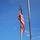 POW/MIA Remembrance Week begins on Peterson Air Force Base The image shows an American flag mounted at the top of a metallic flagpole against a bright blue sky with scattered light clouds. The flag is partially furled and draped around the pole, with its red, white, and blue colors and star-and-stripes pattern clearly visible. A second bare flagpole is visible to the right side of the frame.