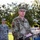 POW/MIA Remembrance Week begins on Peterson Air Force Base A U.S. Air Force officer in OCP (Operational Camouflage Pattern) uniform is speaking at a podium with a microphone at an outdoor military ceremony. A second airman in ABU (Airman Battle Uniform) stands at attention in the background. The setting appears to be a well-maintained military base grounds with trees and grass.