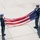POW/MIA week concludes with retreat ceremony Two U.S. Air Force personnel in dress blue uniforms are handling an American flag at a flagpole, likely performing a flag folding or retirement ceremony. Both individuals are wearing white gloves, consistent with formal military ceremonial protocol. The scene takes place on a concrete plaza with multiple flagpoles visible.