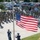 POW/MIA week concludes with retreat ceremony A formal U.S. military flag ceremony is taking place outdoors at what appears to be a military installation. Three honor guard members in dress blue uniforms are raising the American flag on a flagpole while a large formation of military personnel in camouflage uniforms stand at attention and salute. The scene suggests a formal retreat or reveille ceremony with a large assembled audience.