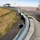 Construction underway for traffic signal outside of Peterson AFB's east gate The image shows a severely damaged and deformed metal guardrail along a highway shoulder, consistent with a vehicle impact. The guardrail has been crushed, bent, and partially torn from its mounting, with sections folded and crumpled along a concrete curb. Orange traffic cones and vehicles are visible in the background, suggesting an active incident or road work zone.