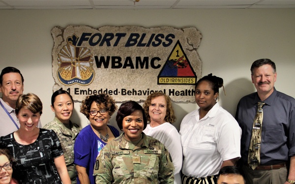 A group photo of approximately 13 military and civilian personnel posing together in an office setting at Fort Bliss WBAMC (William Beaumont Army Medical Center). The sign behind them identifies the unit as '1-1 Embedded Behavioral Health,' part of the 1st Armored Division ('Old Ironsides'). The group includes both uniformed U.S. Army soldiers in ACU/OCP camouflage and civilian staff members.