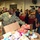 A U.S. Navy sailor named Clifton, wearing digital camouflage NWU (Navy Working Uniform), is sorting through donated food items at what appears to be a food bank or community service event. A second individual wearing a Navy polo shirt observes nearby, while numerous other volunteers work in the background. The setting is a large indoor facility filled with cardboard boxes, food donations, and banana crates.