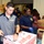 A group of volunteers, several wearing U.S. Navy apparel, are working at what appears to be a food bank or community service event. They are sorting and packing various food items including snacks, peanut butter, and produce into cardboard boxes. The setting is a warehouse-style facility with multiple workers collaborating on the task.