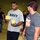 A group of volunteers are working inside what appears to be a food bank or donation center. A man wearing a 'NAVY' t-shirt is holding food boxes while conversing with another man wearing an 'I'm a Hunger Fighter' shirt. Multiple other volunteers are visible in the background handling food items and supplies.