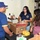 Three individuals are engaged in what appears to be a food drive or donation sorting activity at a table covered with cardboard boxes and various food items. A woman wearing a navy blue polo shirt with an official emblem is actively handling items from a Coca-Cola branded cardboard box. The setting appears to be a storage or utility room with tools and equipment visible in the background.
