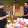 Two individuals are working together at what appears to be a food bank or donation sorting facility. One person wearing a black t-shirt and blue cap is holding a food item while the other, wearing a dark uniform shirt with an official patch, holds an open cardboard box. They are surrounded by large quantities of donated food items, boxes, and packaged goods.