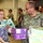 A U.S. Navy service member in digital camouflage uniform works alongside a young civilian woman in a tie-dye shirt at what appears to be a food bank or donation distribution center. They are handling boxes of food products, including Annie's Organic Cookie Bites and Goldfish crackers. Several other volunteers and personnel are visible working in the background of the warehouse-style facility.