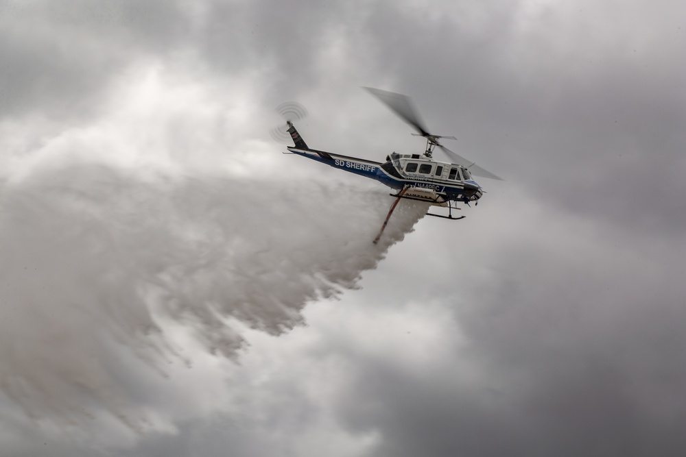 A San Diego Sheriff's Department helicopter is performing a water or fire retardant drop against an overcast sky. The aircraft, a Bell 205/UH-1 type helicopter marked 'SD SHERIFF', is releasing a large cascade of white liquid from its belly tank. This appears to be a firefighting operation, likely in response to a wildfire in the San Diego County region.