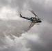 A San Diego Sheriff's Department helicopter is performing a water or fire retardant drop against an overcast sky. The aircraft, a Bell 205/UH-1 type helicopter marked 'SD SHERIFF', is releasing a large cascade of white liquid from its belly tank. This appears to be a firefighting operation, likely in response to a wildfire in the San Diego County region.
