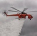 An Erickson Air-Crane helicopter (number 729) is captured mid-flight performing a water drop operation against an overcast, grey sky. The bright orange heavy-lift helicopter is releasing a large cascade of water from its belly tank, indicating active aerial firefighting operations. The image is sharp and well-composed, clearly showing the aircraft's identifying markings and the dramatic water release.