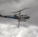 A San Diego County Sheriff's helicopter is captured in flight against an overcast, cloudy sky. The blue and white helicopter, registration N549RL, is equipped with a firefighting water/retardant tank suspended beneath its fuselage. The image is sharp and well-composed, clearly showing the aircraft's markings and external equipment.