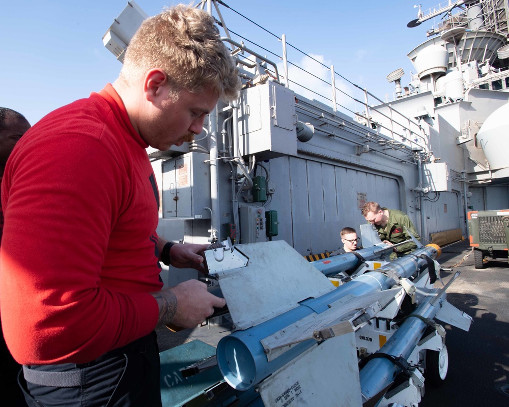 Military personnel are working on what appears to be ordnance or missile equipment on the deck of a naval vessel. Three individuals are visible, with one in a red shirt in the foreground handling documentation or a checklist near the equipment. The setting is clearly a warship deck with naval superstructure visible in the background.