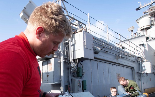 Military personnel are working on what appears to be ordnance or missile equipment on the deck of a naval vessel. Three individuals are visible, with one in a red shirt in the foreground handling documentation or a checklist near the equipment. The setting is clearly a warship deck with naval superstructure visible in the background.