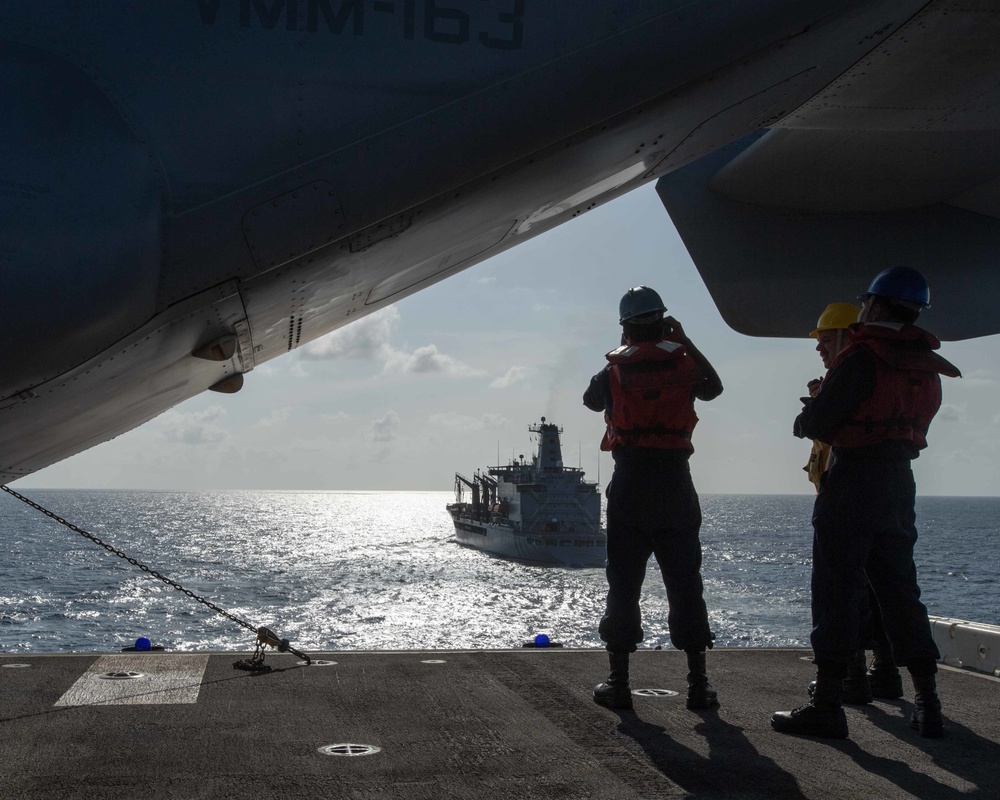 Two naval personnel wearing red life vests and colored hard hats stand on the flight deck of a warship, observing an approaching replenishment vessel at sea. The scene is framed beneath the wing and fuselage of a parked military aircraft, with partial markings visible reading 'VMM-163'. The image captures what appears to be a replenishment-at-sea (RAS) or approach operation in open ocean conditions.