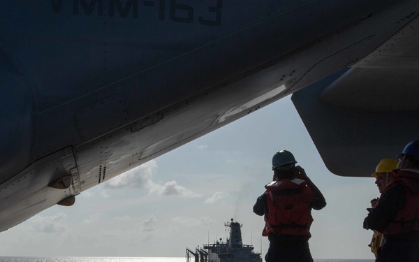Two naval personnel wearing red life vests and colored hard hats stand on the flight deck of a warship, observing an approaching replenishment vessel at sea. The scene is framed beneath the wing and fuselage of a parked military aircraft, with partial markings visible reading 'VMM-163'. The image captures what appears to be a replenishment-at-sea (RAS) or approach operation in open ocean conditions.