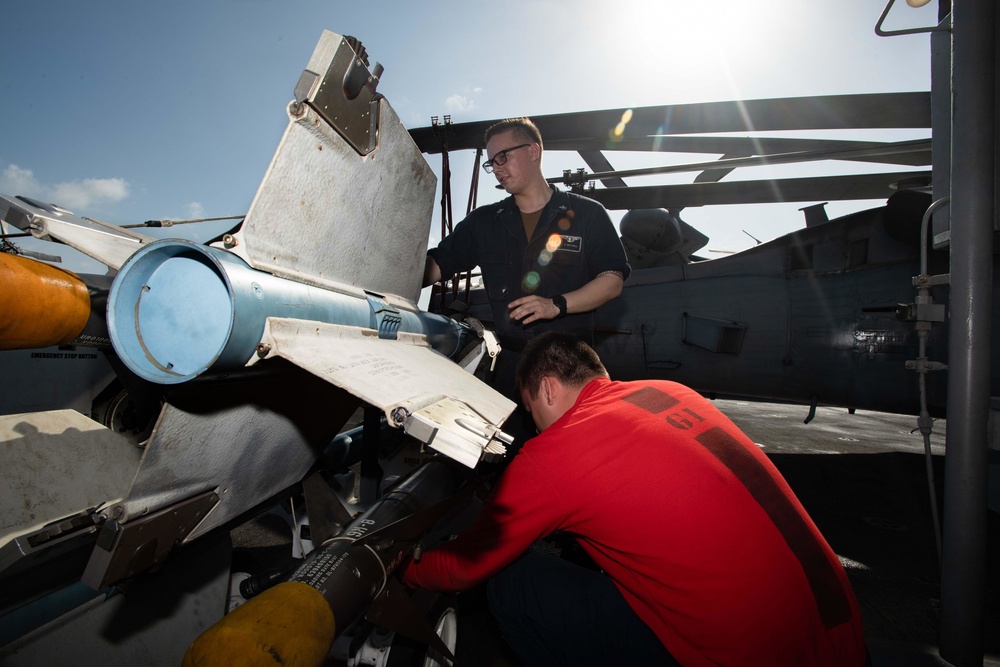 Two military personnel are performing maintenance or weapons loading work on a helicopter, likely aboard a naval vessel. One individual wearing a dark uniform with glasses is observing, while another in a red shirt is actively working on what appears to be a weapons system or mechanical component. The setting appears to be an outdoor flight deck under bright sunlight.
