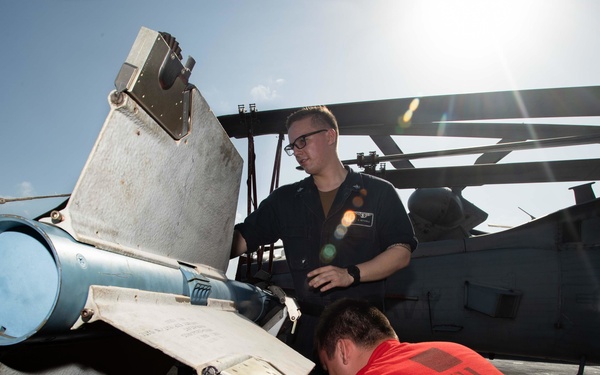 Two military personnel are performing maintenance or weapons loading work on a helicopter, likely aboard a naval vessel. One individual wearing a dark uniform with glasses is observing, while another in a red shirt is actively working on what appears to be a weapons system or mechanical component. The setting appears to be an outdoor flight deck under bright sunlight.