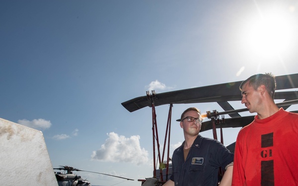 Three military personnel are working on what appears to be weapons or ordnance mounted on a naval aircraft, likely on the deck of an aircraft carrier or naval vessel. Two individuals are wearing red long-sleeve shirts with visible markings, while a third wears a blue naval uniform with glasses. The scene takes place outdoors under bright sunlight with a partially cloudy sky.