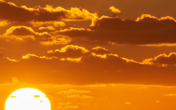 A dramatic silhouette of a large naval vessel, likely an aircraft carrier, is photographed against a vivid orange and golden sunset over open ocean. The large solar disk is positioned just behind the ship, creating a striking backlit composition. The sky is filled with scattered clouds illuminated in deep orange and amber tones.