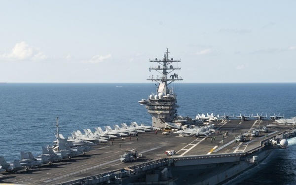 A large US Navy nuclear-powered aircraft carrier is underway at sea, photographed from an elevated aerial perspective. The flight deck is populated with numerous fixed-wing aircraft arranged in organized rows. The vessel is moving at speed, as evidenced by the visible wake trailing behind the stern.